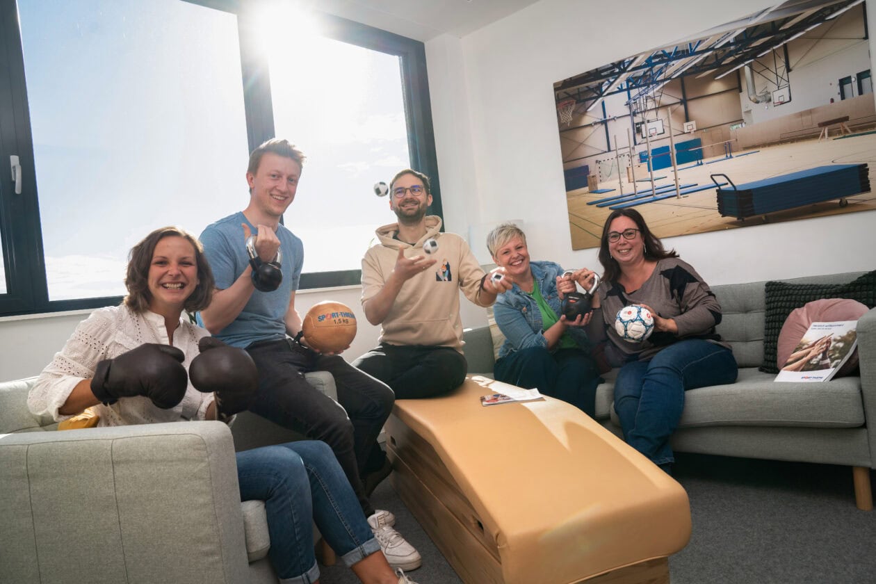 Fünf Mitarbeitende aus der Personalabteilung von Sport-Thieme sitzen in einem Besprechungsraum und halten verschiedene Sportgeräte in der Hand.