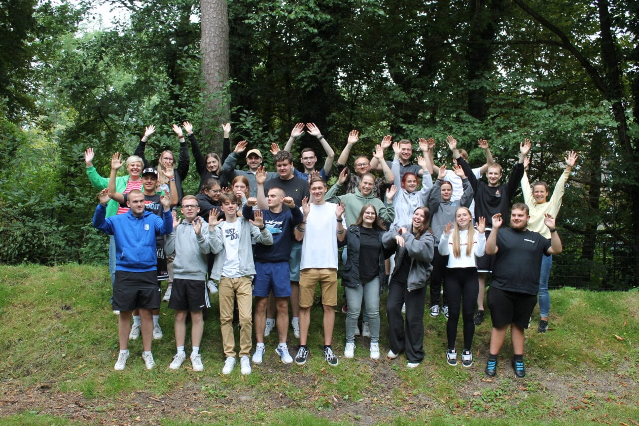 A group of Sport-Thieme apprentices stands outdoors, waving at the camera, surrounded by trees.