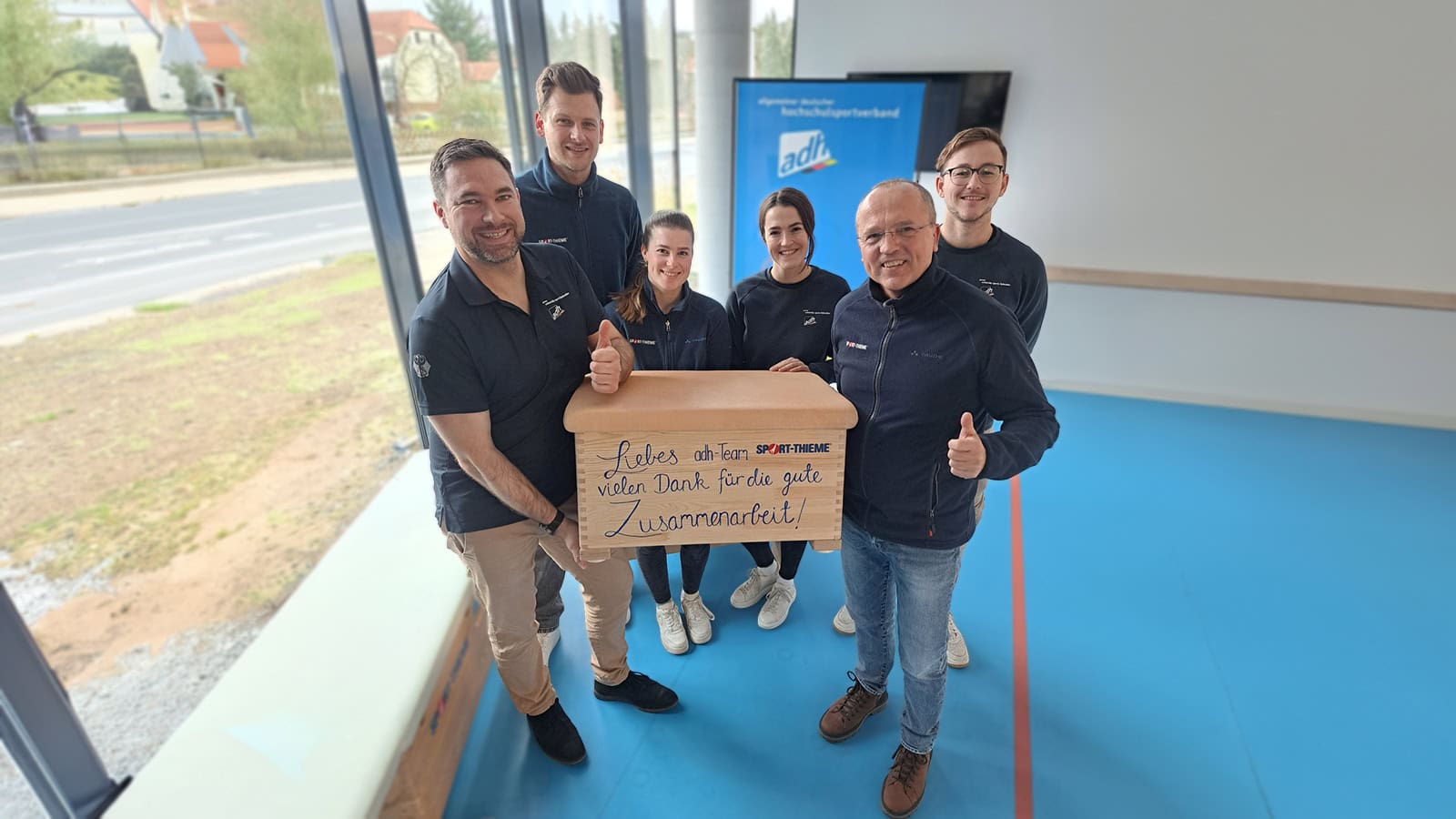 Employees of Sport-Thieme and the adh hold a gymnastics box together, smiling while looking at the camera.