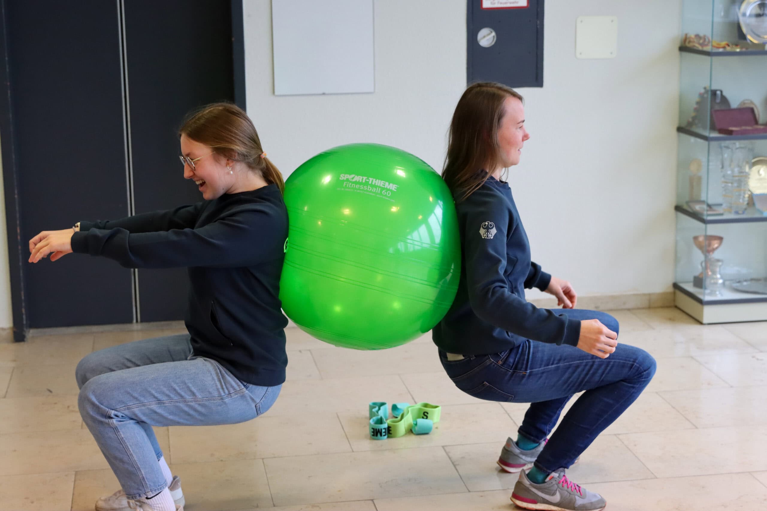 Two people perform an exercise together with a green fitness ball, sitting in a squat position.