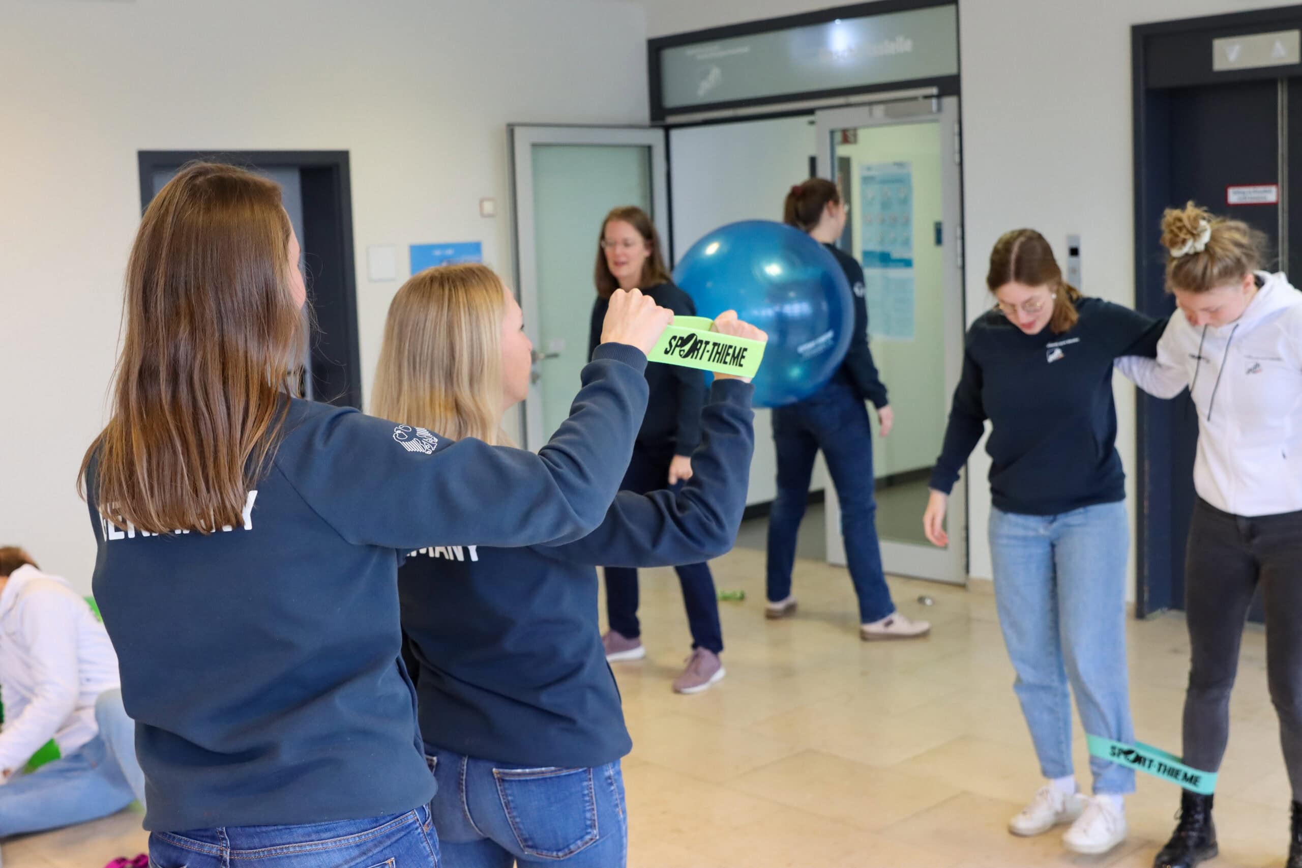 Two people hold a Sport-Thieme band while others in the background are actively using a ball and other materials.