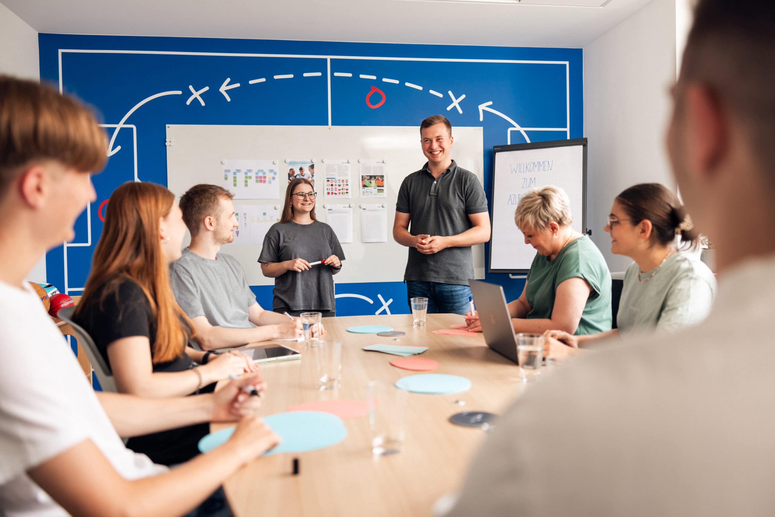 Sport-Thieme trainees and mentors in a meeting room. Two people stand at the front leading the trainee meeting.