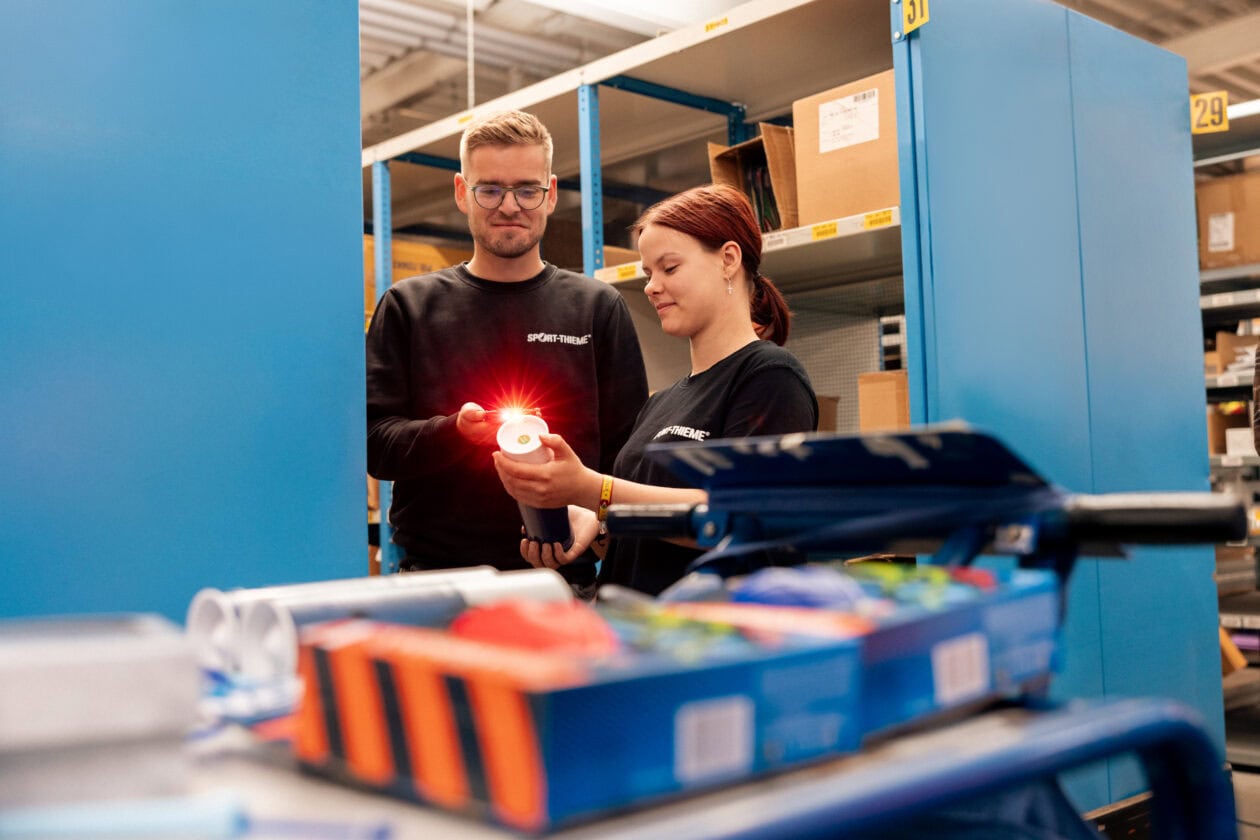 Two Sport-Thieme employees stand in front of a warehouse shelf, scanning an item.
