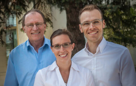 The management team of Sport-Thieme, consisting of three people in white and blue shirts, stands in front of a tree outdoors.