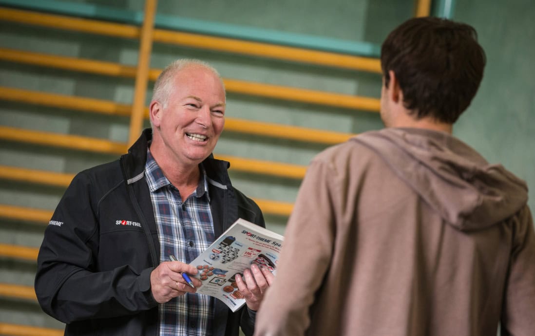 A Sport-Thieme employee is advising another person in a gym, holding an informational brochure.