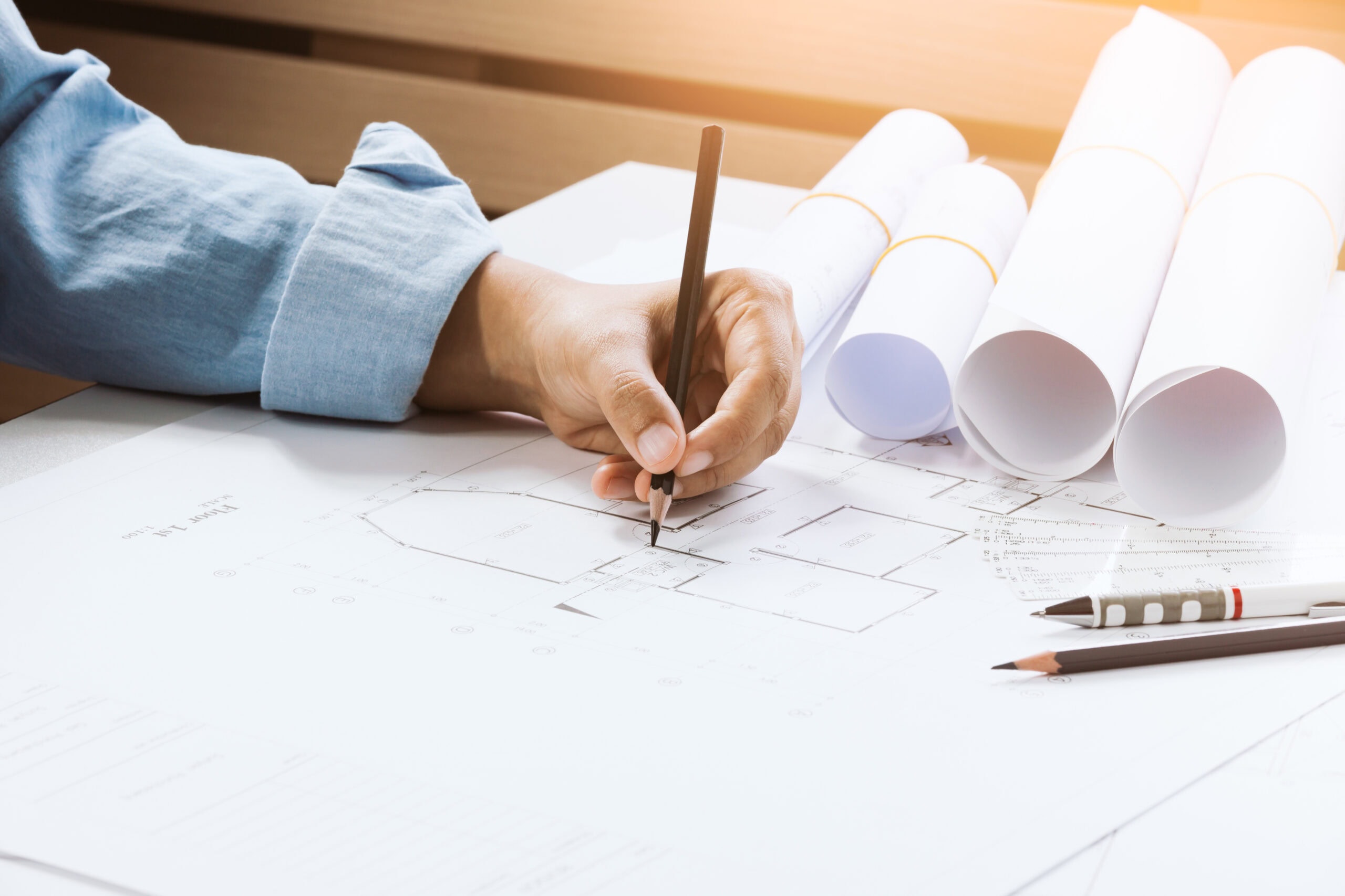 A person drawing a plan with a pencil, surrounded by paper rolls and pens on a table.