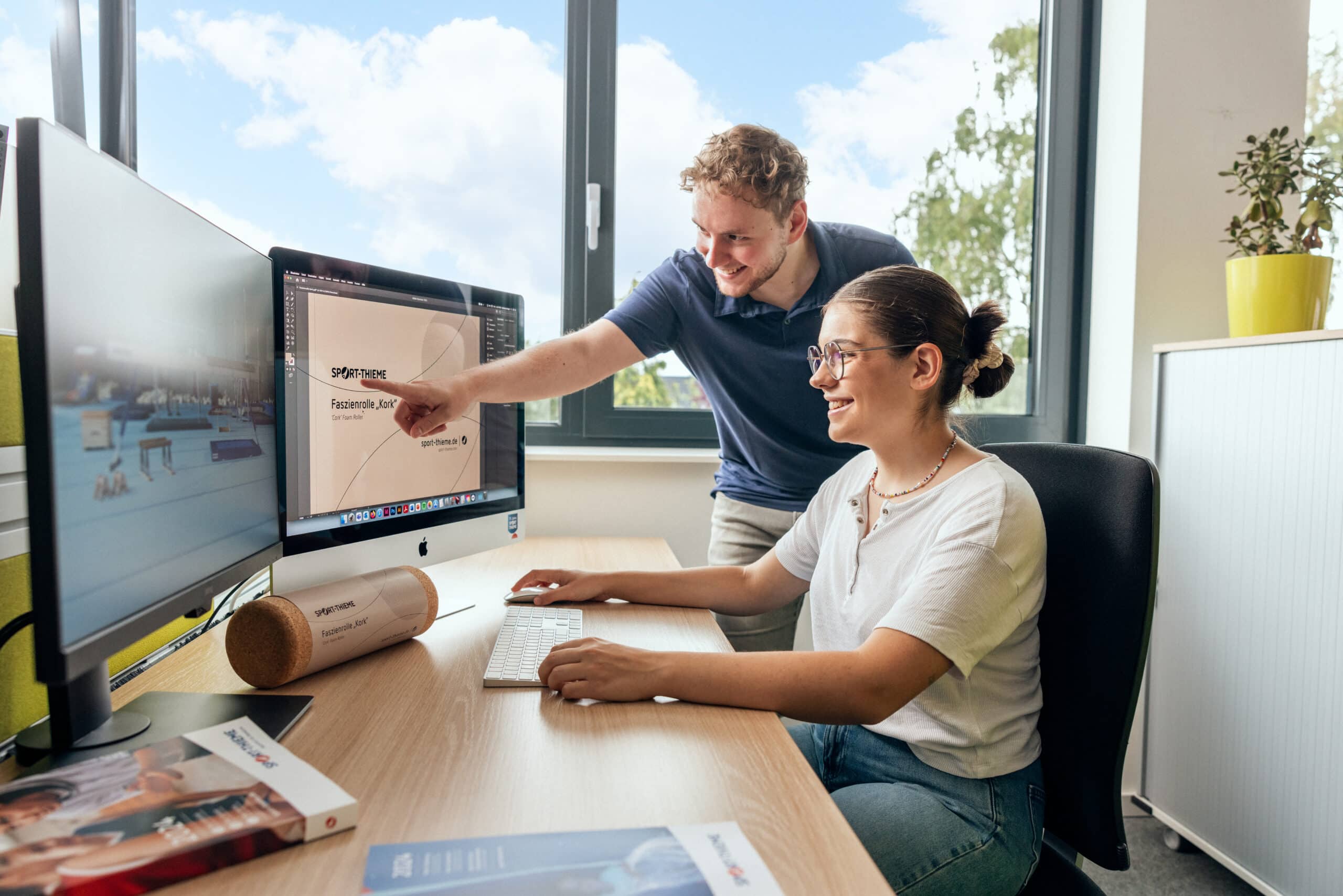 Azubi in der Mediengestaltung arbeitet an einem Produktdesign mit Unterstützung eines Kollegen.