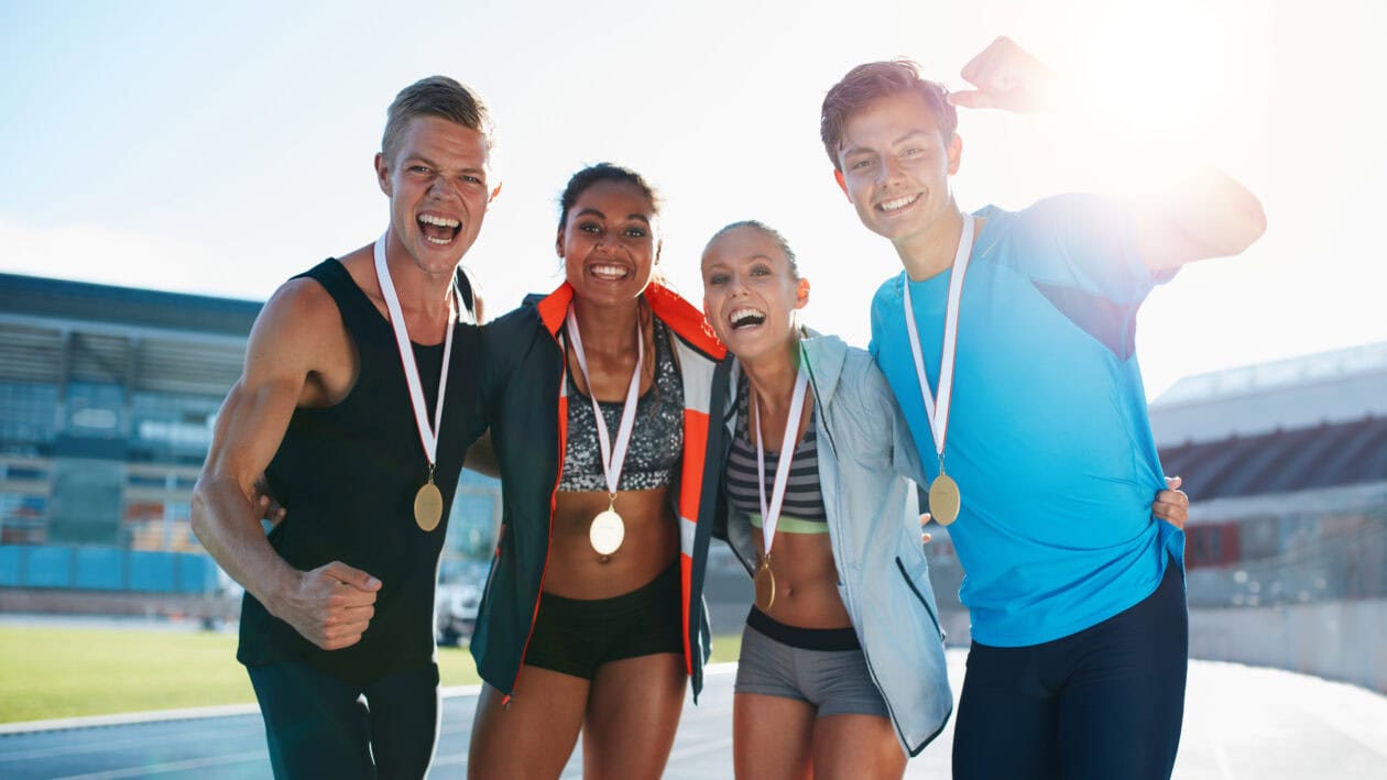 Four athletes wearing medals celebrate together in the stadium.
