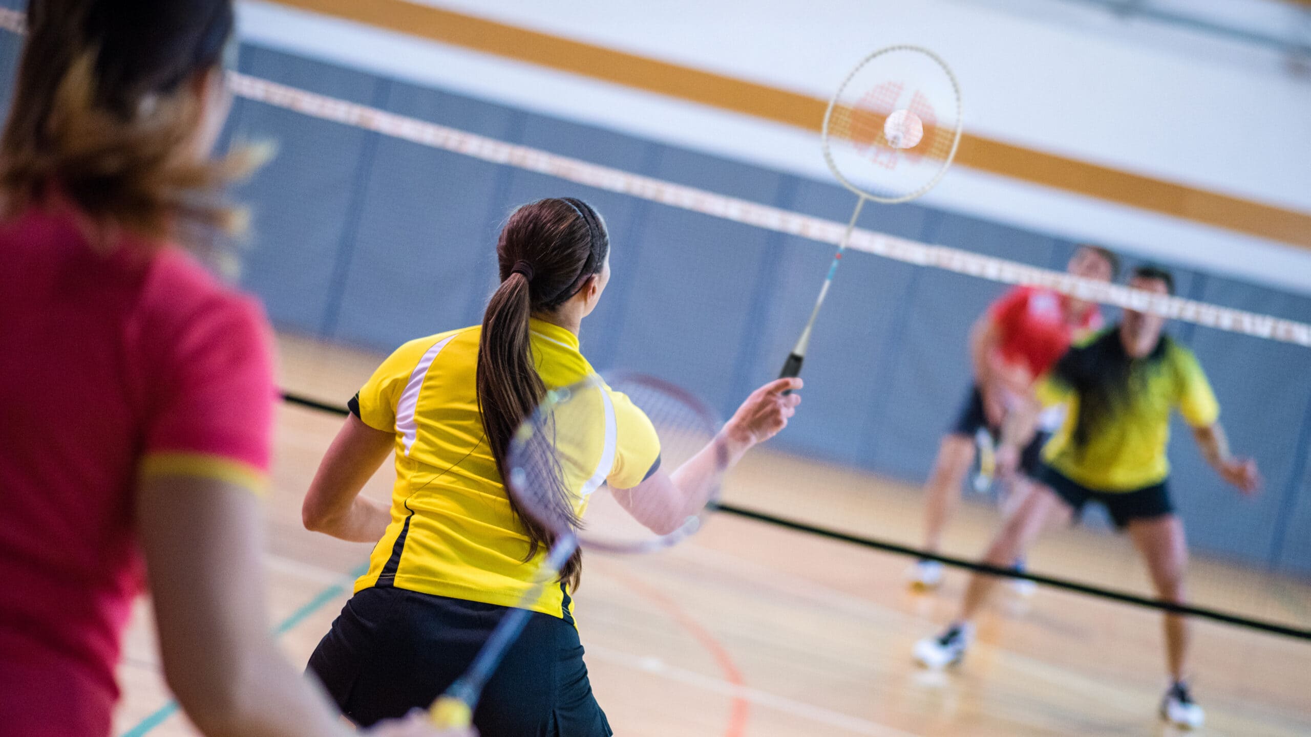 Four people play doubles badminton in a gym.