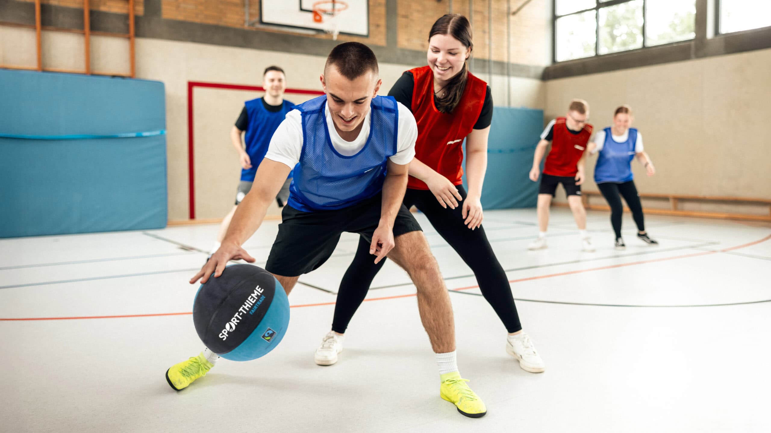 Young people play basketball indoors wearing colored bibs.