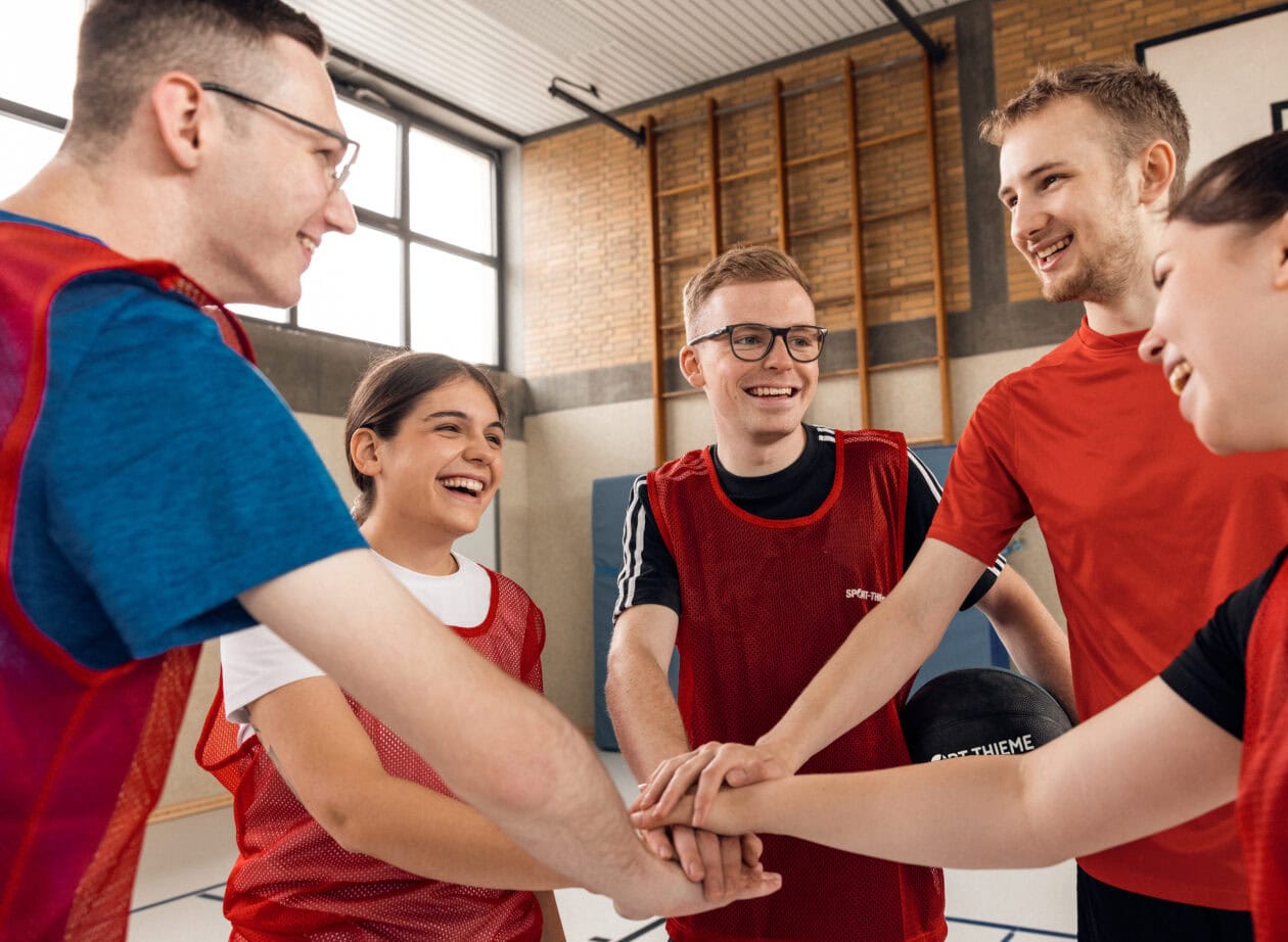 Sport-Thieme trainees in red jerseys standing in a circle, placing hands together and laughing.