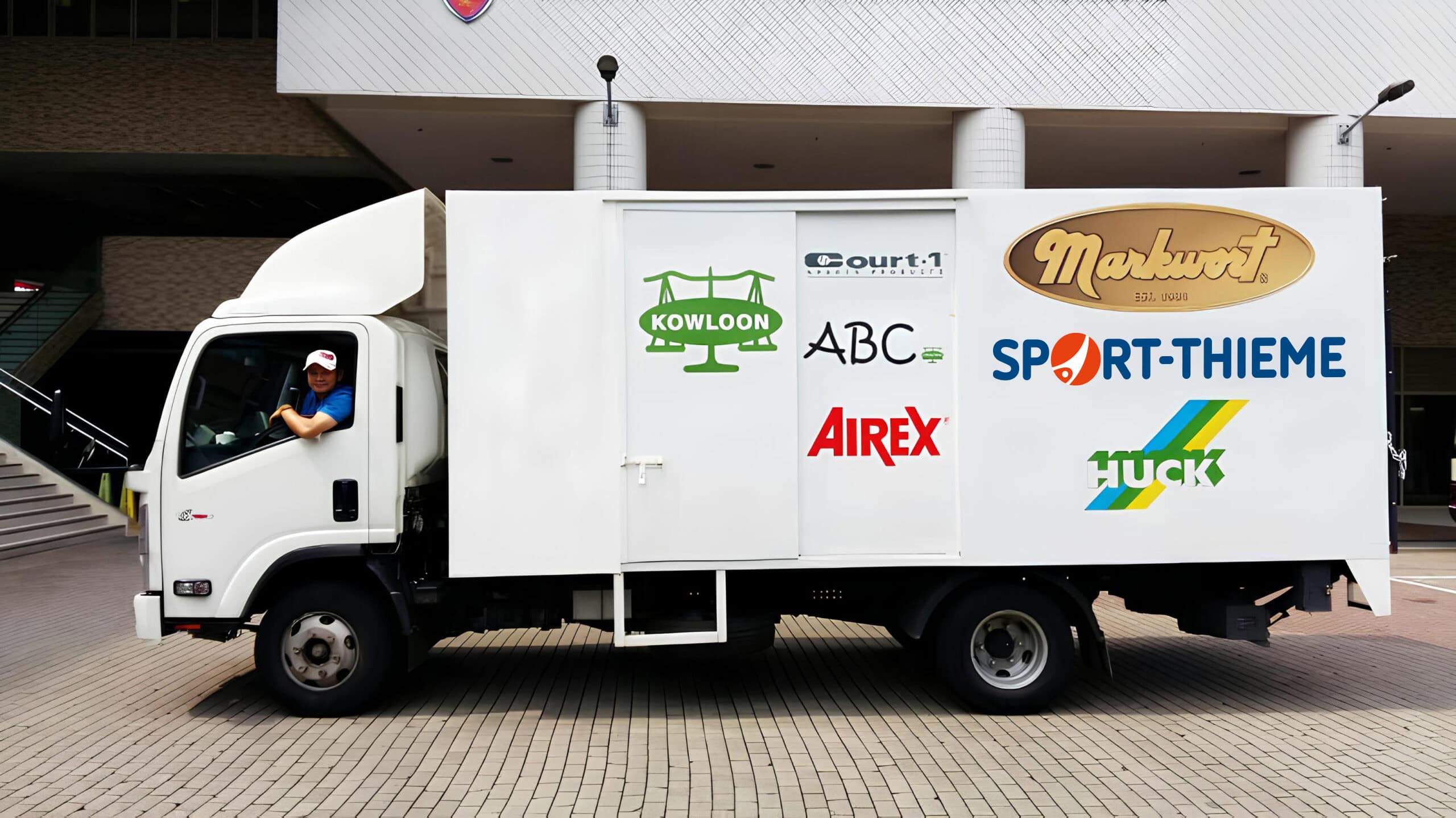 A truck with logos of Sport-Thieme and other brands is parked in front of a building, with the driver looking out the window.