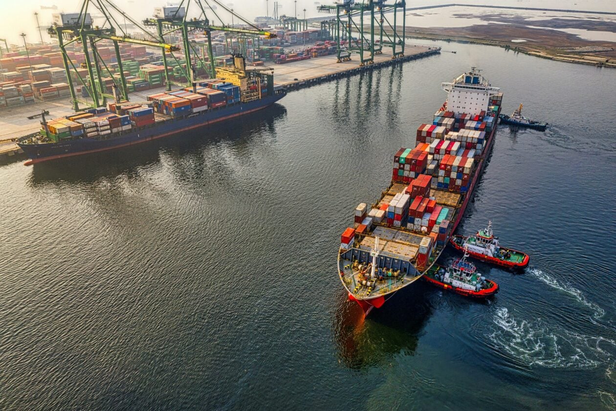 A large container ship is being maneuvered in the port by two small tugboats, while additional cargo ships and container cranes can be seen in the background.